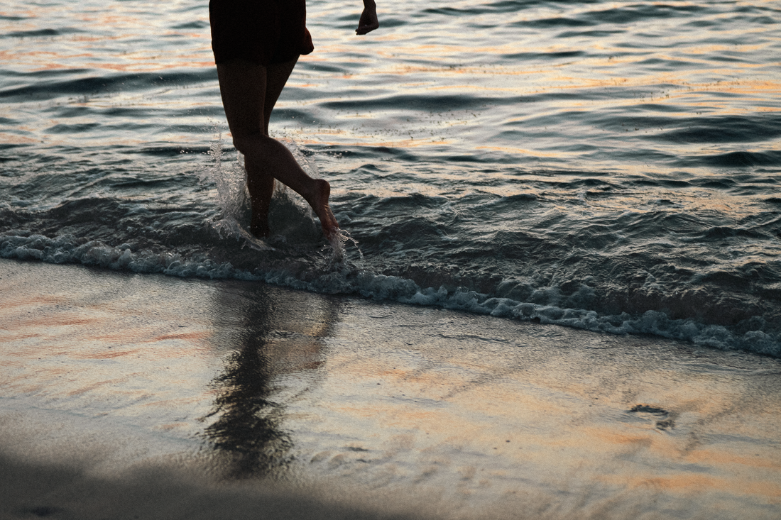 Person running barefoot through shallow ocean waves at sunset, with reflections on wet sand.